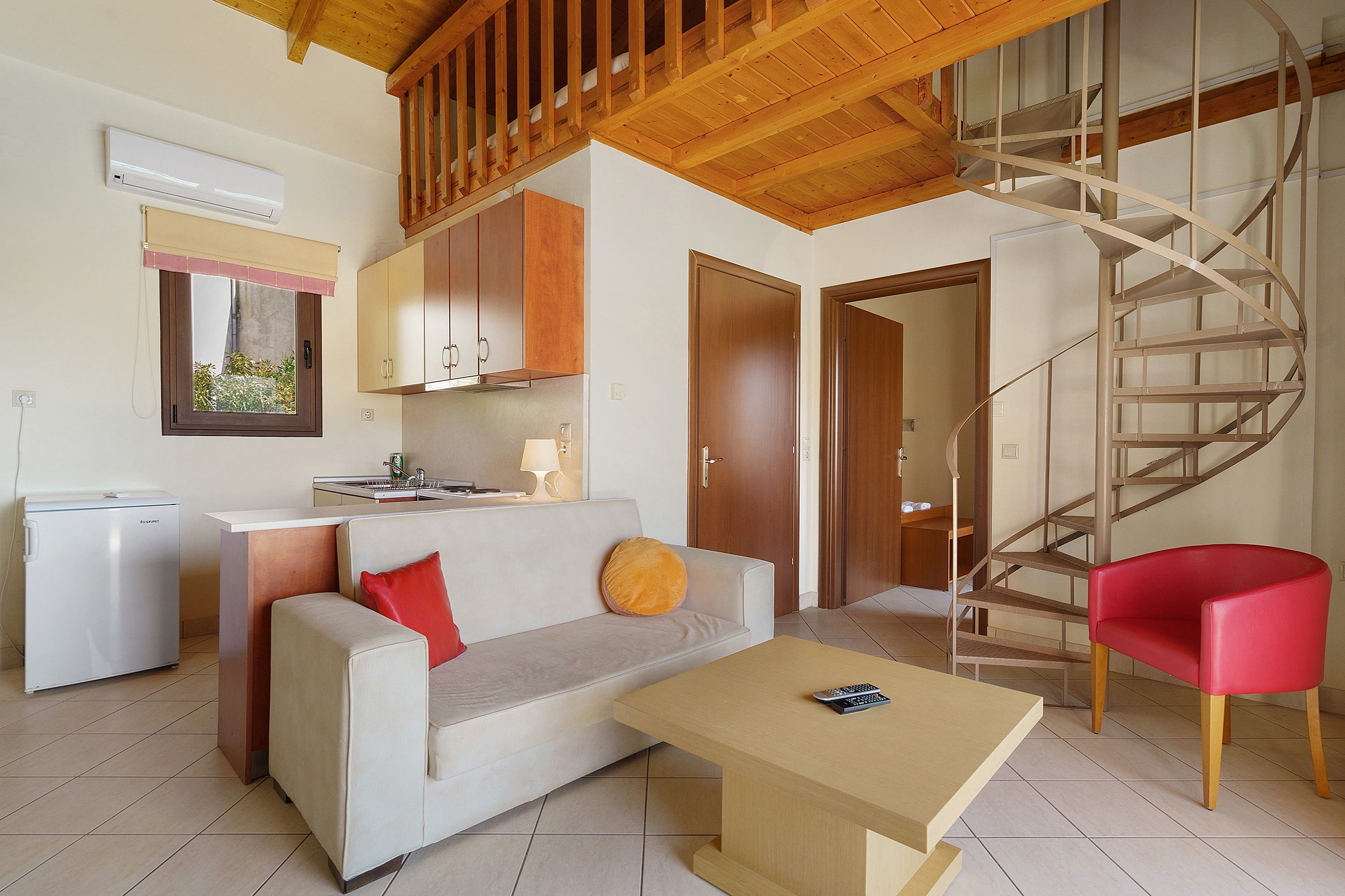 Un espacio habitable compacto con kitchenette, un sofá, una mesa de centro, una silla roja y una escalera de caracol.