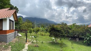 A house with a red-tiled roof and a brick exterior, surrounded by a lush green garden with a stone pathway leading to the entrance. - Cozy La Chorrera Cabin Surrounded by Nature with a View of the Mountains (Venecia)