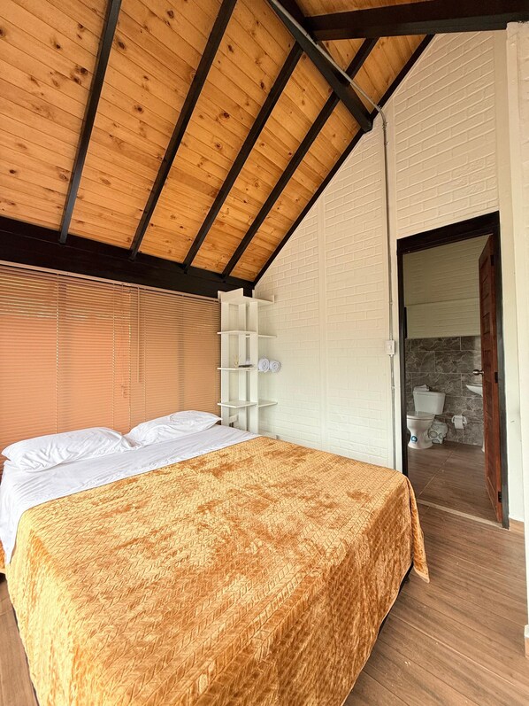 A bedroom with a wooden ceiling, a bed with a patterned bedspread, a white wall with a brick pattern, and a bathroom visible through an open door.