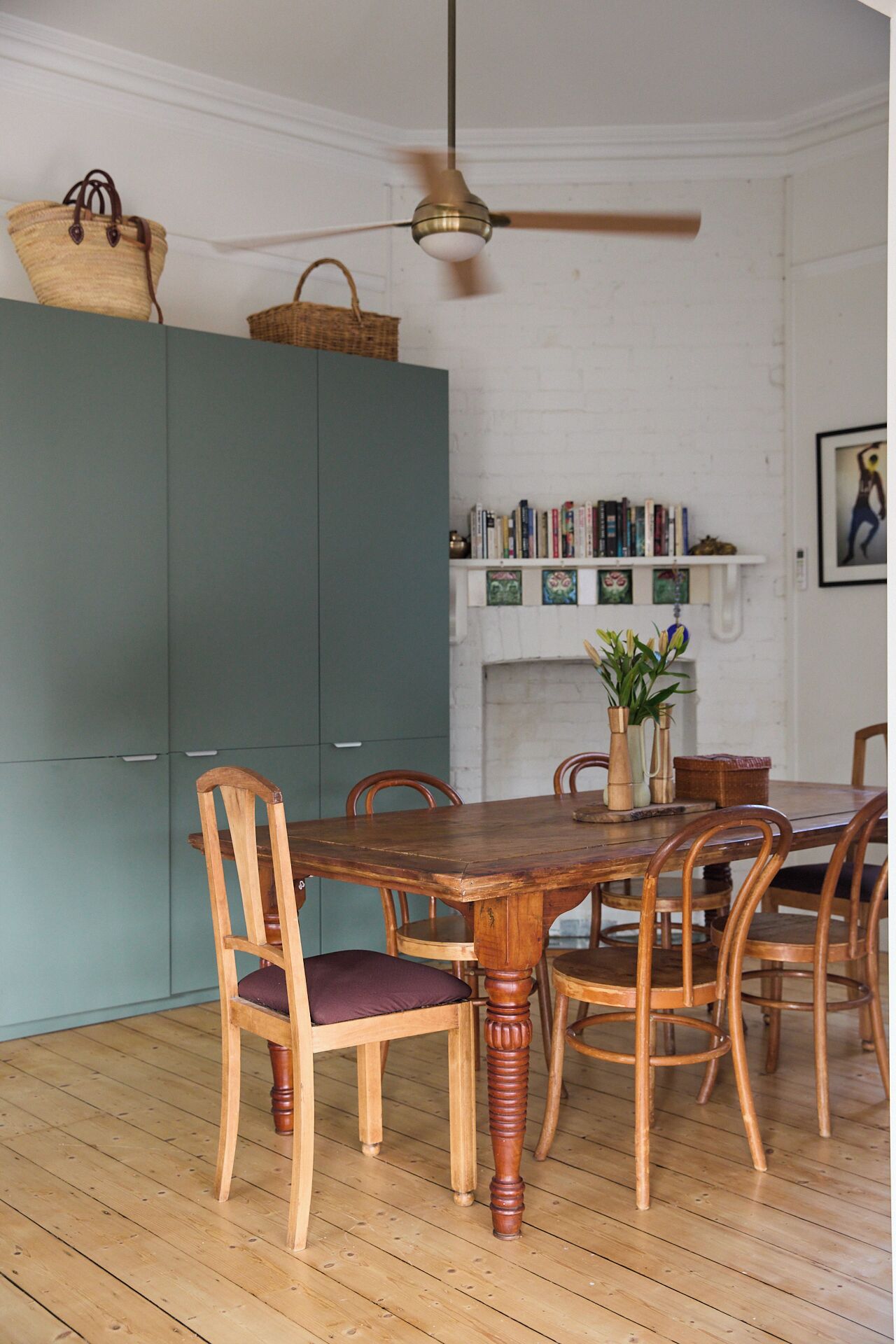 A dining area with a wooden table and chairs, a ceiling fan, and a bookshelf against a white wall.