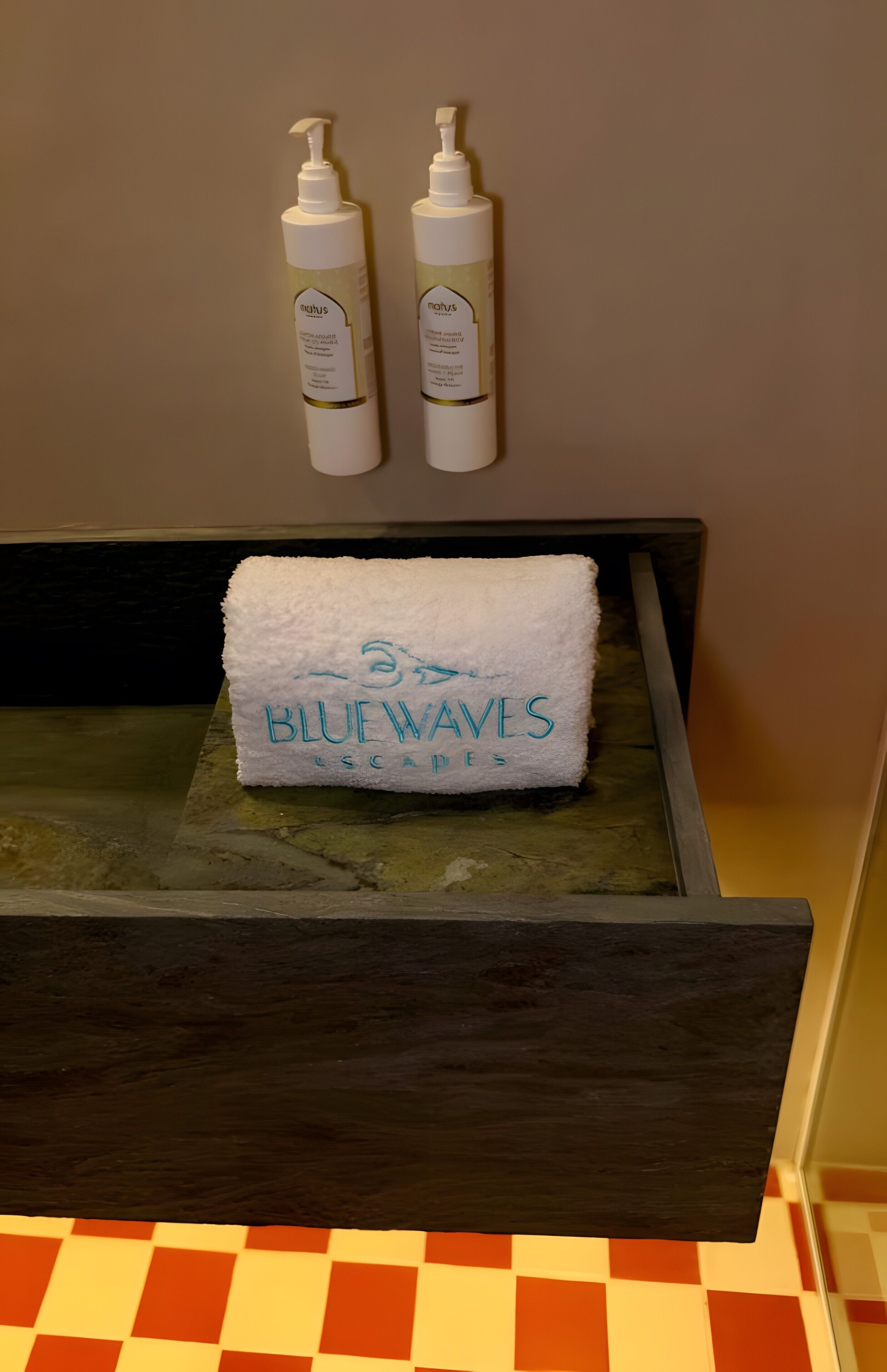 Et badeværelse med et håndklædestativ, der holder to flasker og et håndklæde med teksten 'BLUE WAVES'.