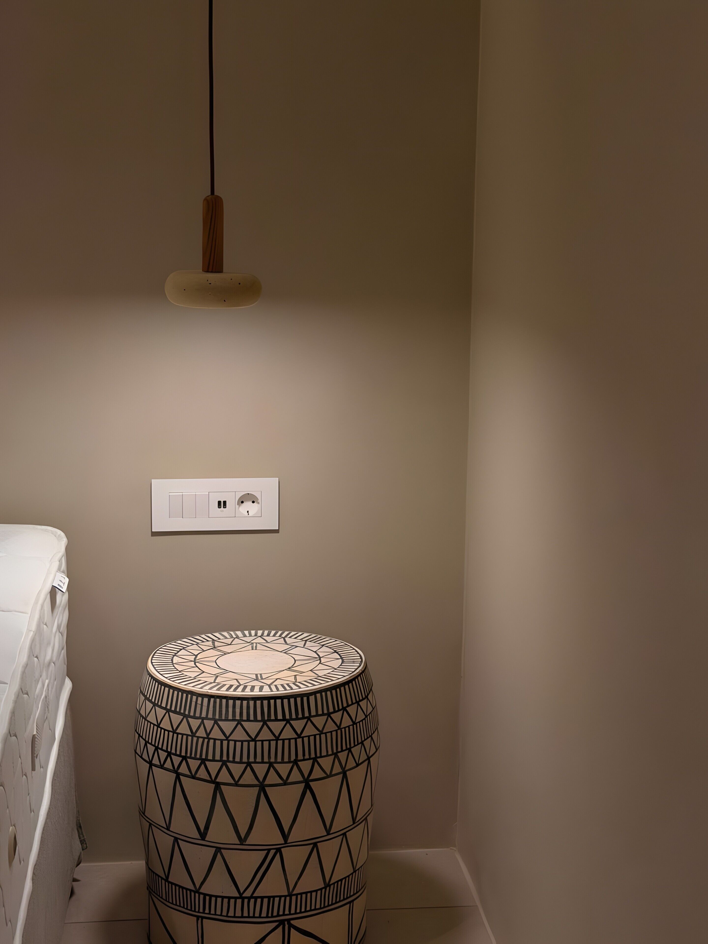 Ein Zimmer mit einer weißen Matratze, einem rundem Tisch mit Muster, einer Pendelleuchte und einer Steckdose.