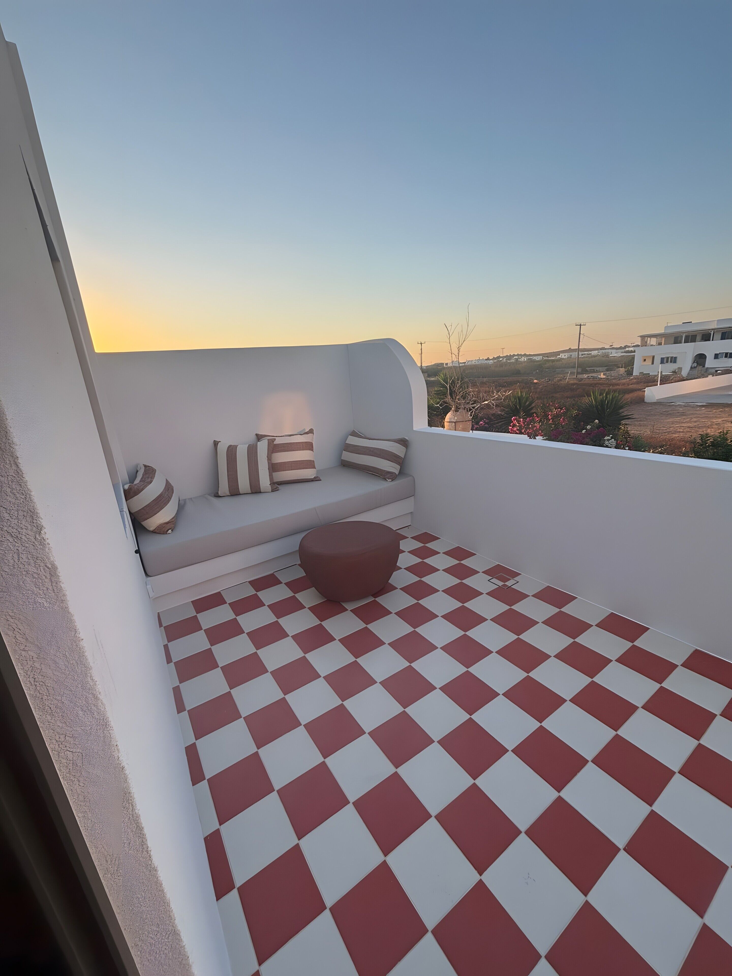 Een balkon met een zwart-wit geruit vloertje, een zithoek met gestreepte kussens, en uitzicht op een gebouw en het omliggende landschap.