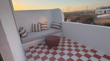 Een balkon met een zwart-wit geruit vloertje, een zithoek met gestreepte kussens, en uitzicht op een gebouw en het omliggende landschap.