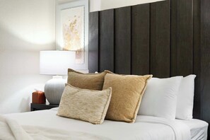 Una cama con ropa blanca y dos almohadas color beige, una lámpara de noche y un cuadro en la pared.