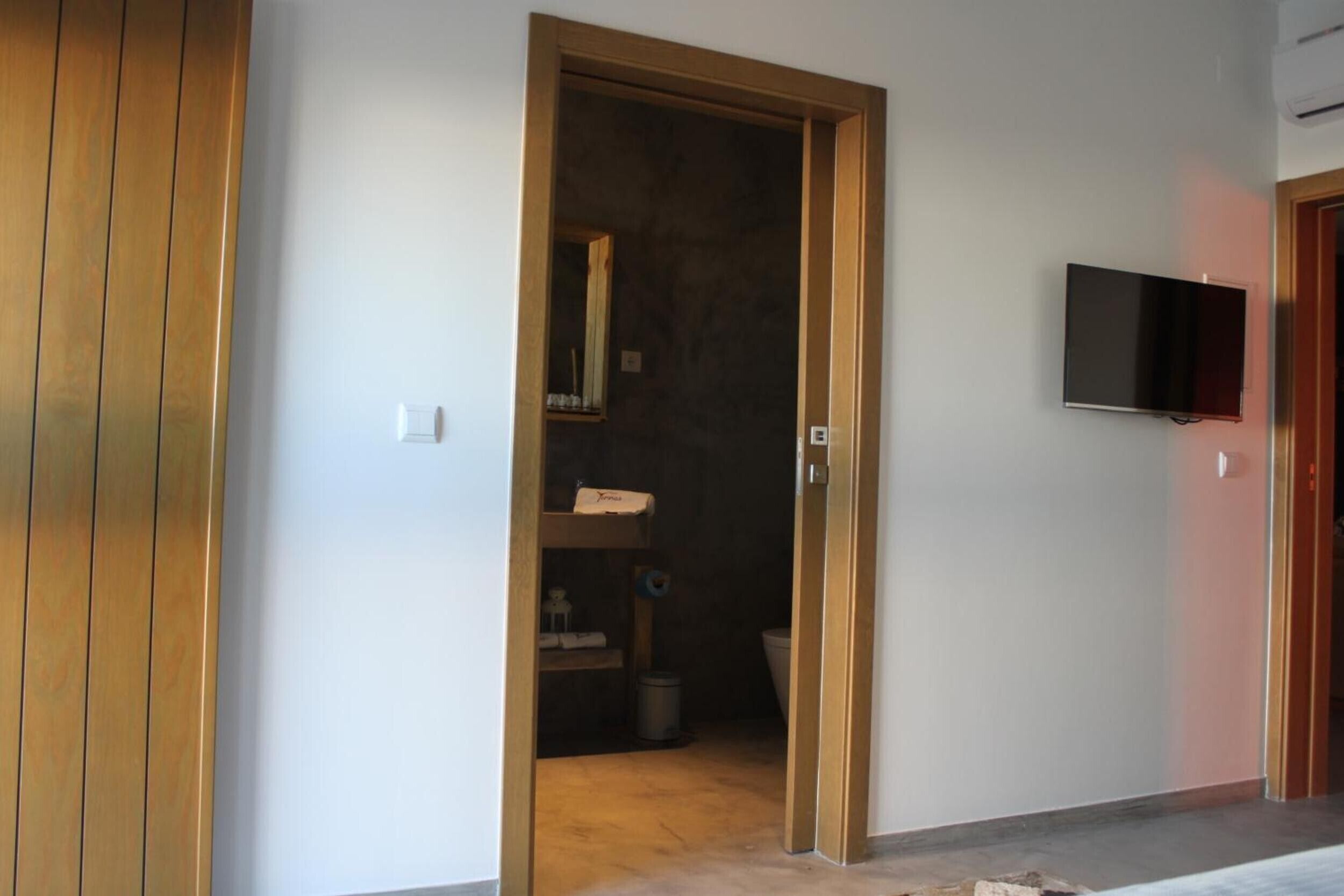 Ein Zimmer mit Holztür, einem an der Wand befestigten Fernseher und einem sichtbaren Badezimmer mit Toilette und Waschbecken.