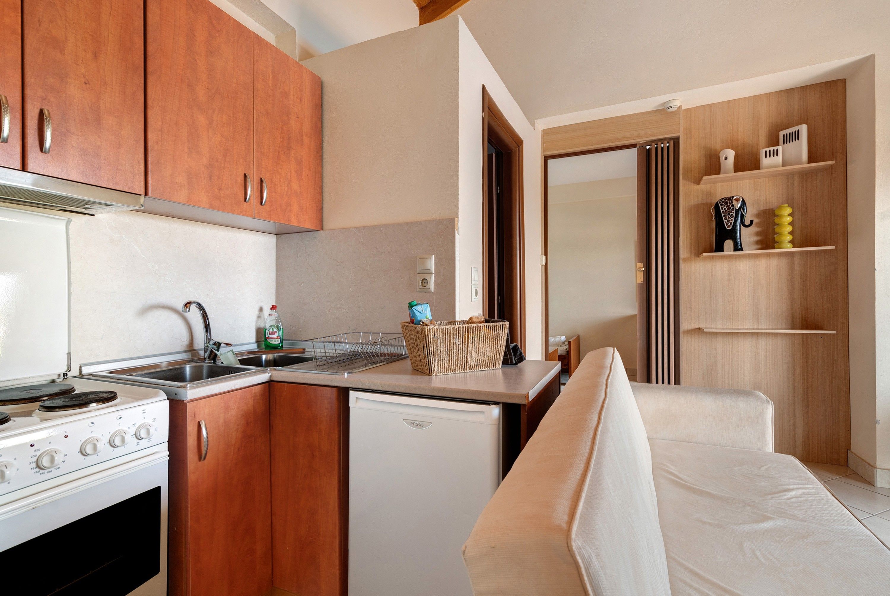 A compact kitchen with a stove, sink, and a small refrigerator. There is a wooden cabinet above the stove and a white countertop.