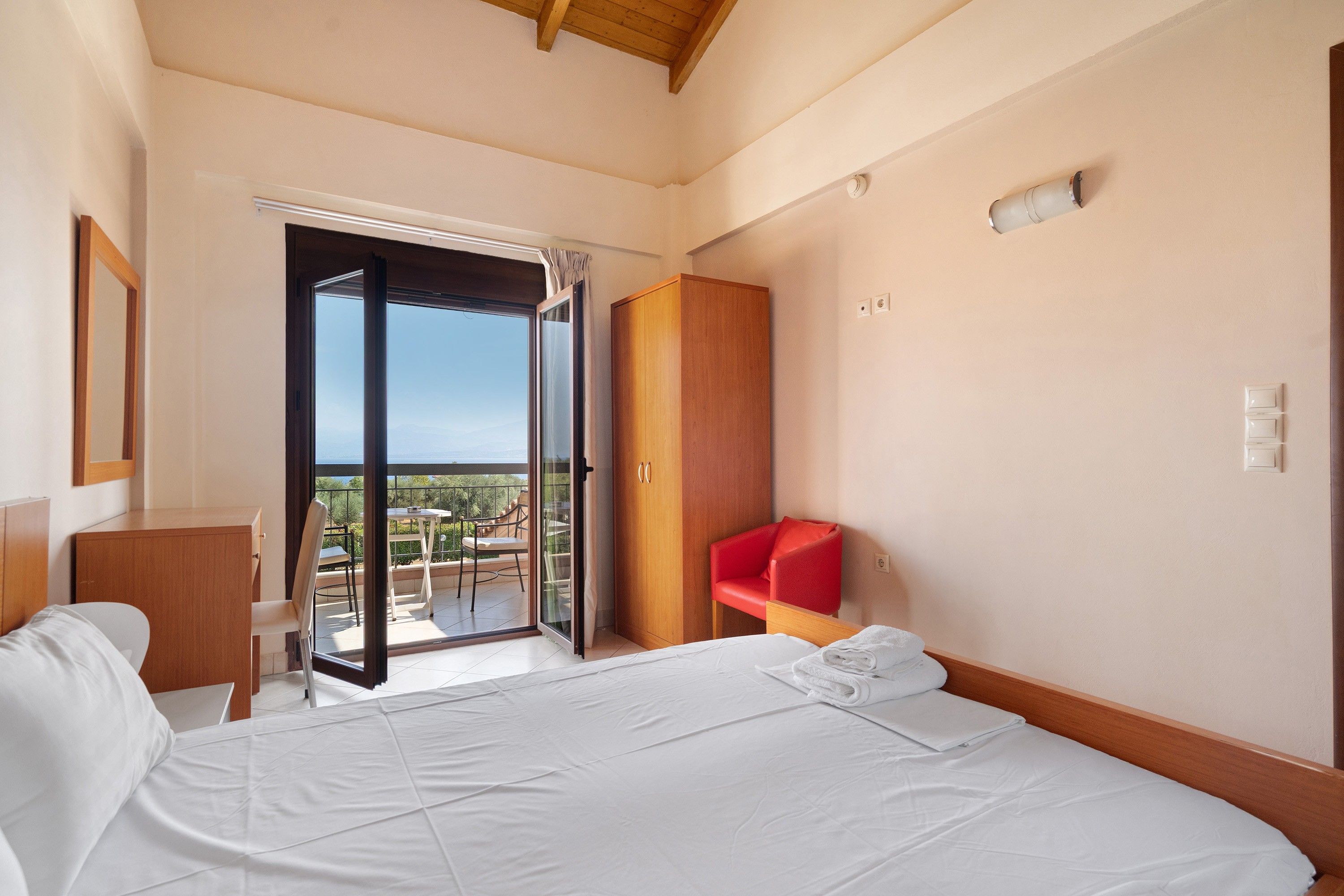 Een hotelkamer met een bed, een rode stoel, een houten garderobe en een schuifdeur met toegang tot een buitenruimte.
