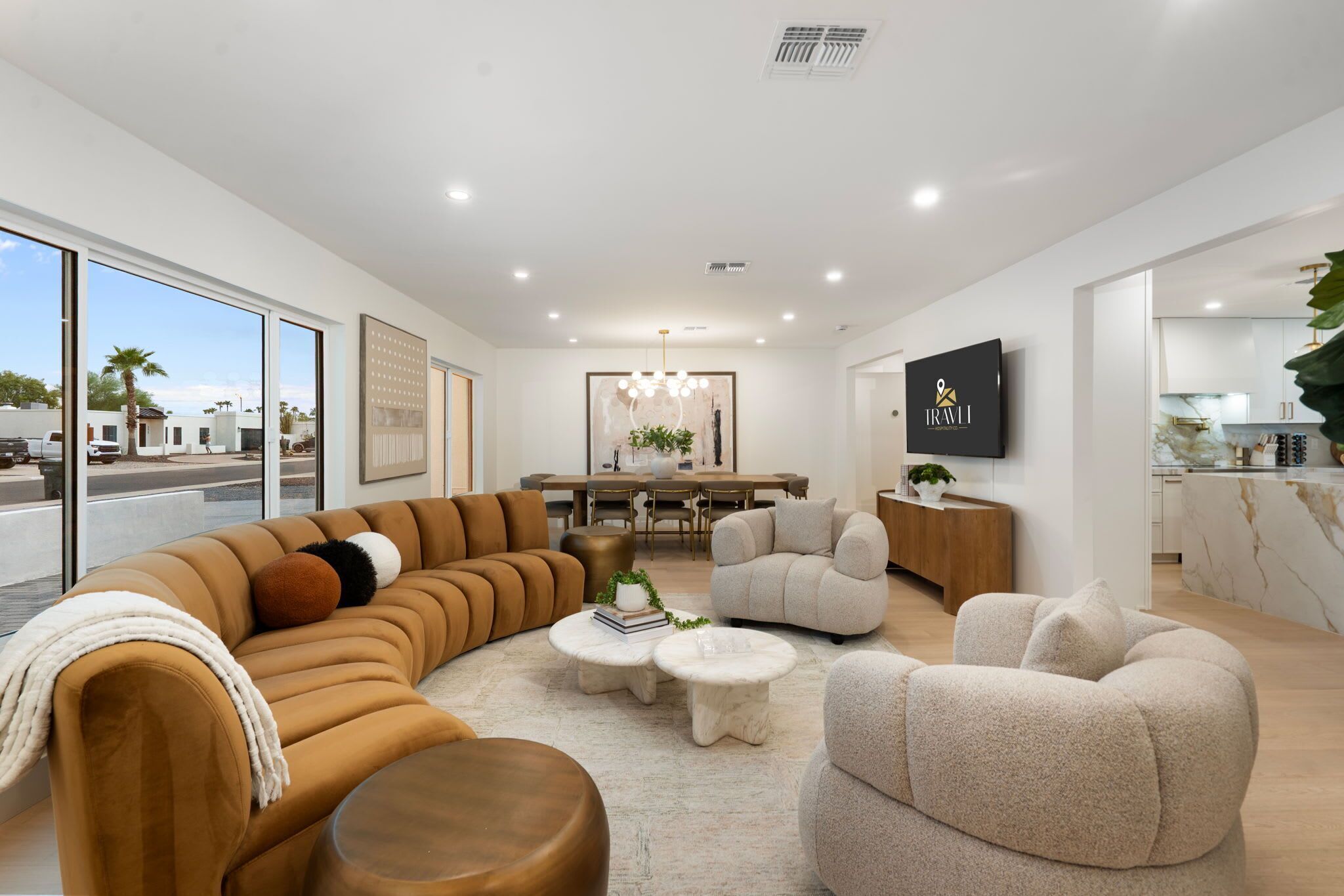 A modern living room with a sectional sofa, round coffee table, and armchairs. There is a dining area with a table and chairs, and a kitchen with a marble countertop.