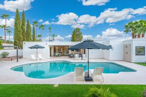 Ein moderner Poolbereich im Freien mit Liegestühlen und Sonnenschirmen, umgeben von viel Grün und Palmen.
