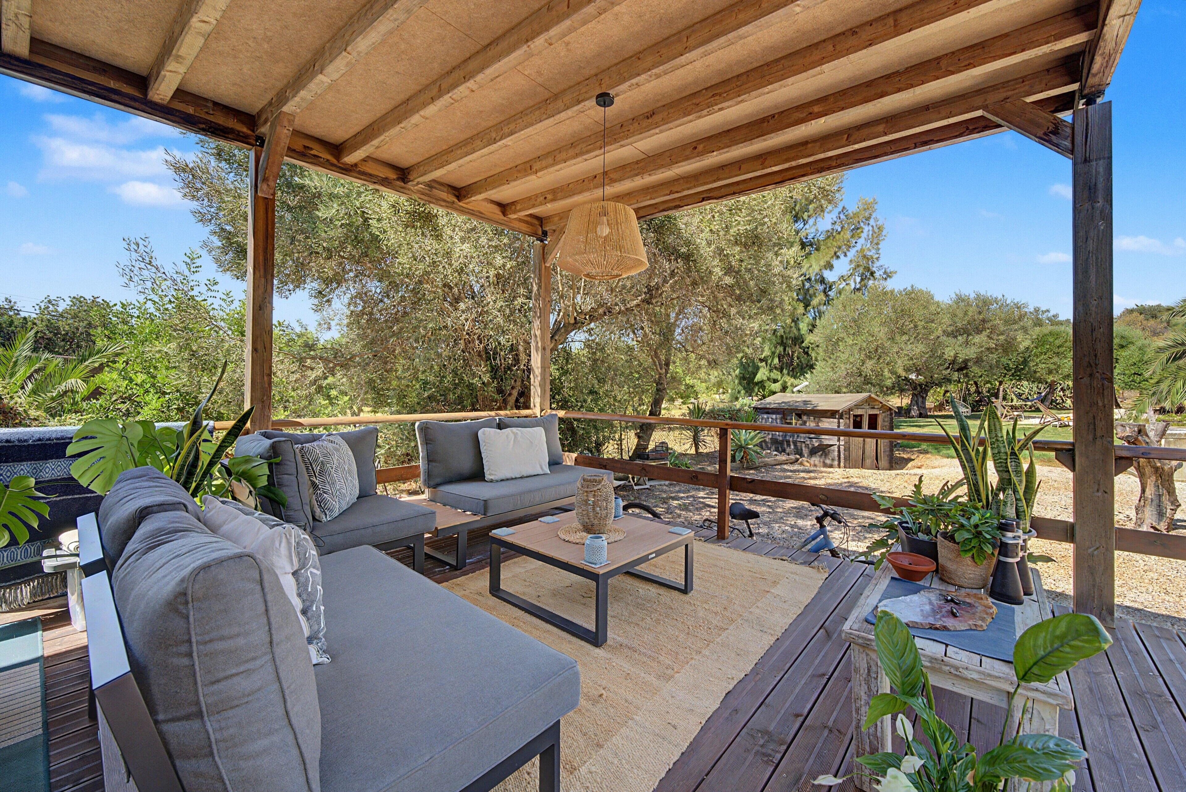 An outdoor patio with a wooden pergola, grey sofas, a coffee table, and various potted plants.