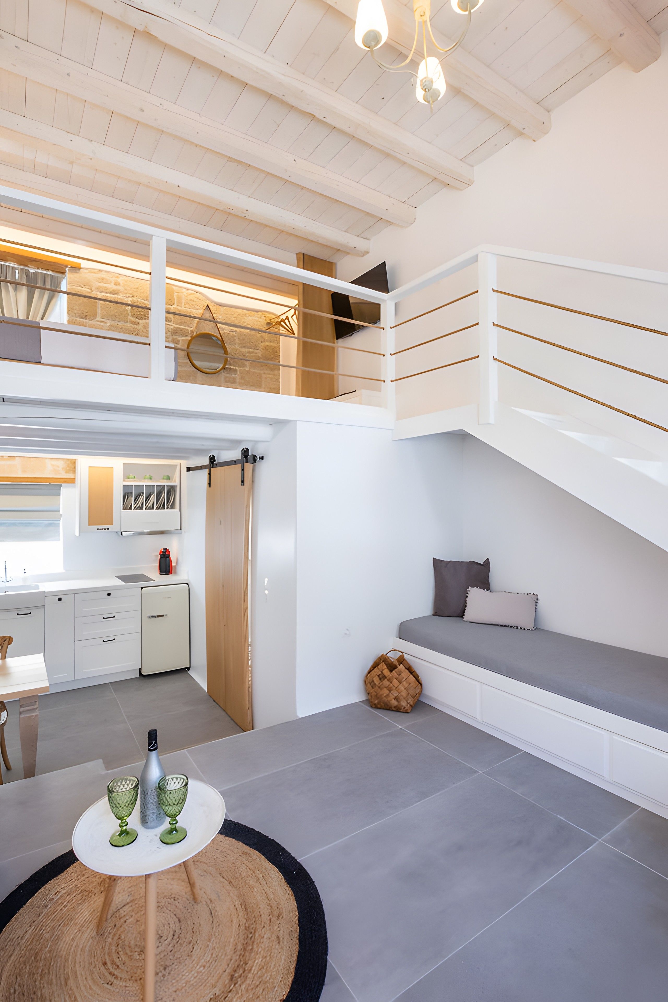 Un dormitorio estilo loft moderno con cama elevadas, una zona de comedor y una cocina al fondo, más una mesa redonda de madera con vasos.