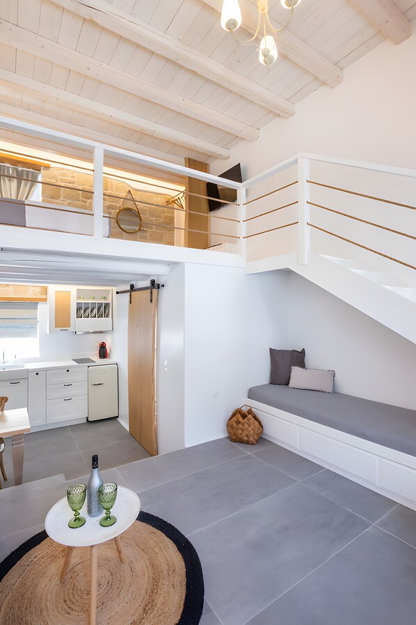 A modern loft bedroom with a loft bed, a dining area with a kitchen in the background, and a round wooden table with glasses.