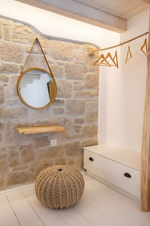 A room with a stone wall, a round mirror, a wooden shelf, a hanging wooden hanger, a woven pouf, and a white dresser. - Villa 'Grannys 1' with Mountain View, Wi-Fi and Air Conditioning (Karpathos)