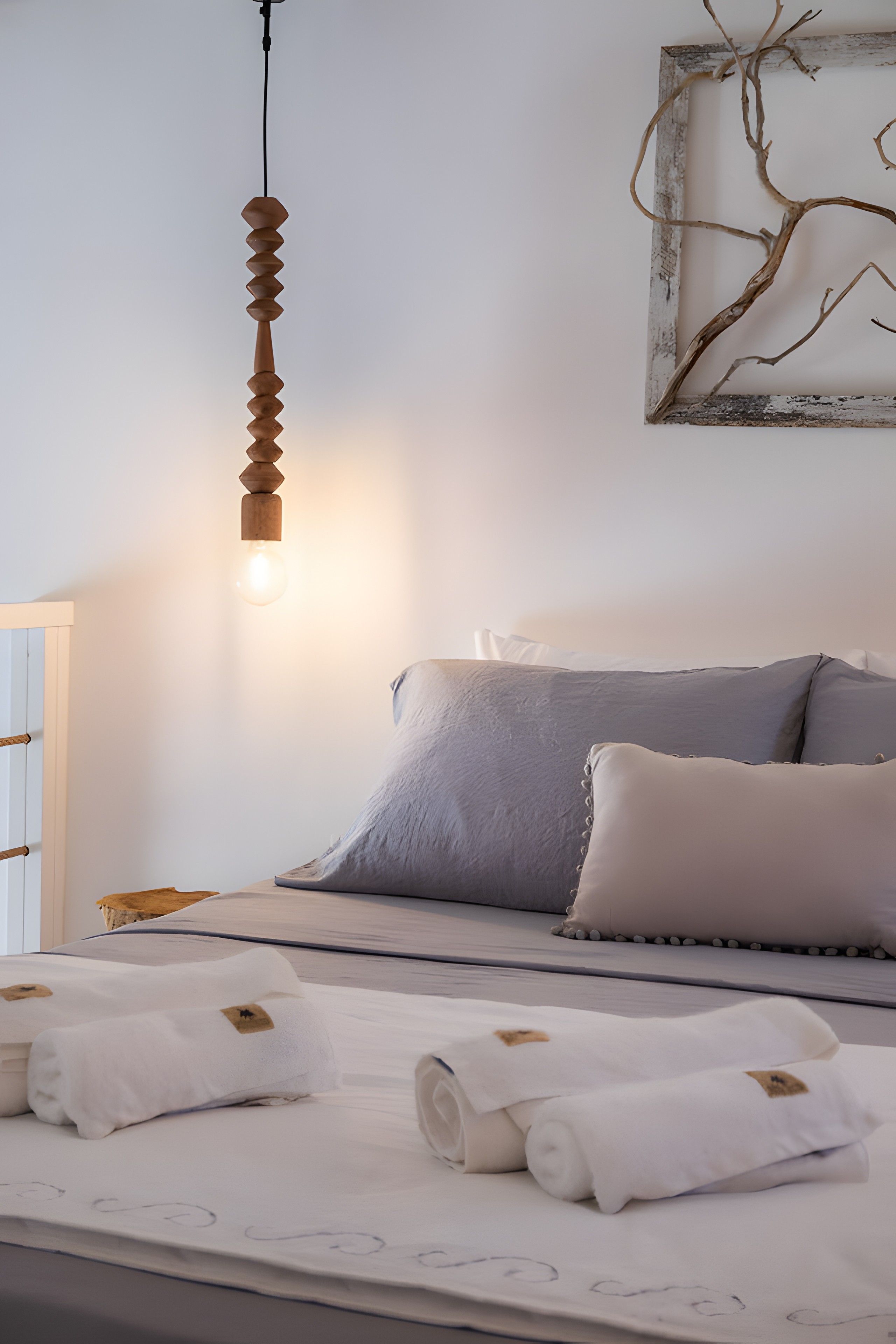 A neatly made bed with pillows and a blanket, a hanging light fixture, and a wall with a branch artwork.