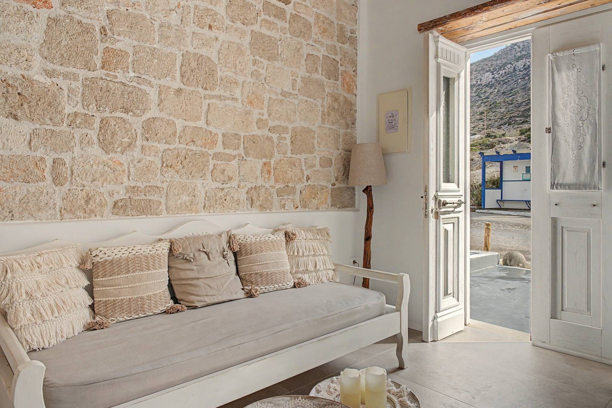 A room with a stone wall, a white sofa with patterned cushions, a door leading outside, and a small table with a glass.