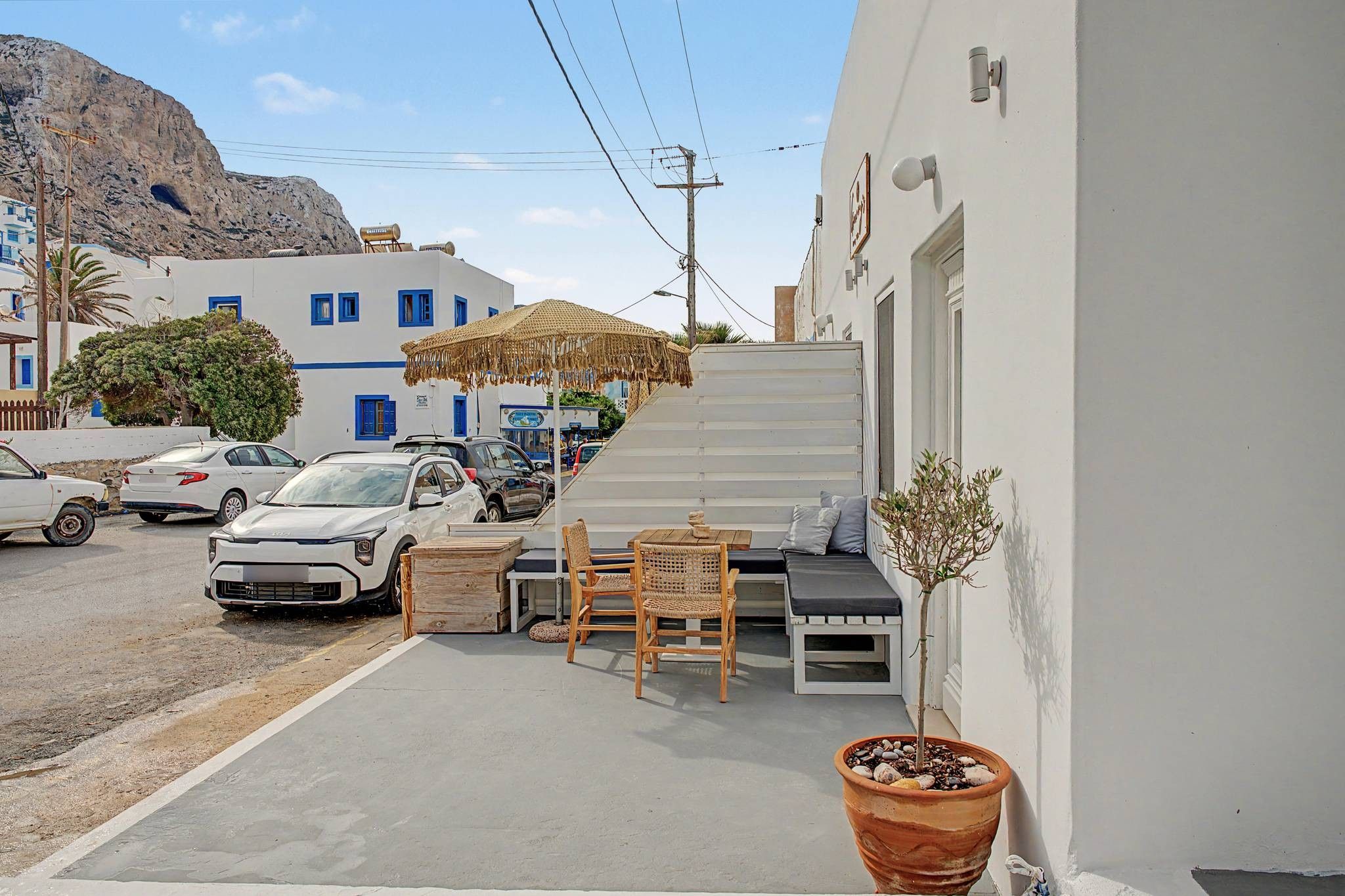 A narrow street with parked cars, a white building with blue shutters, a wooden bench, and a potted plant.
