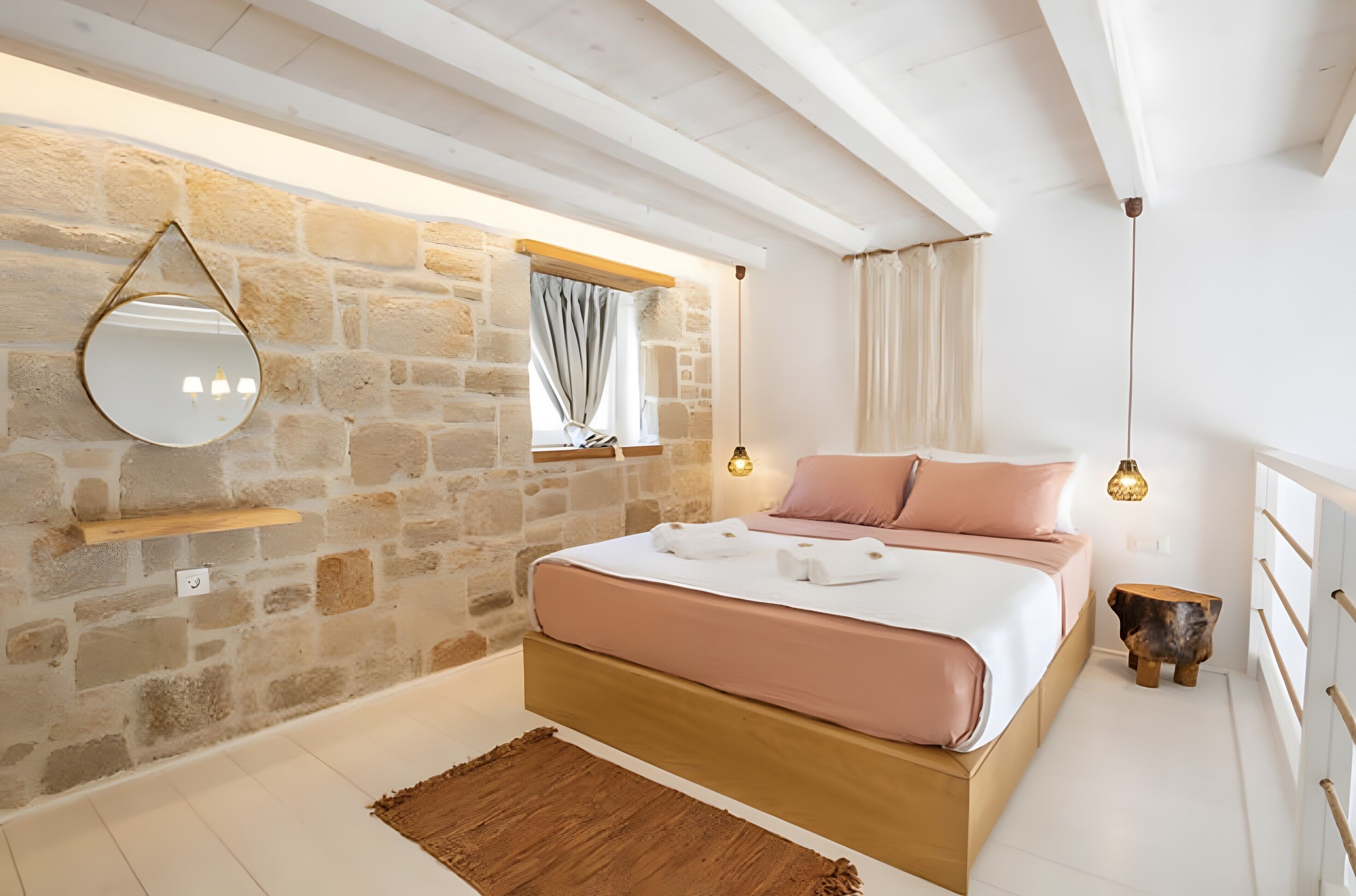 Une chambre avec un mur en pierre, un lit avec une literie rose, une tête de lit en bois, un luminaire suspendu et un miroir.