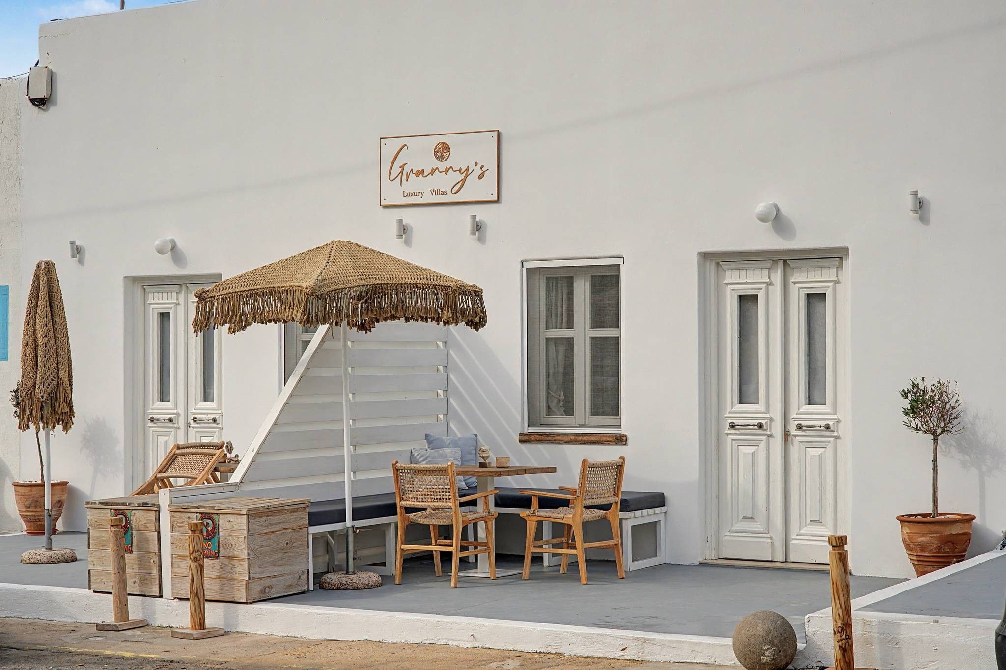 Un edificio blanco con un letrero que dice 'Granny's Luxury Villas'. Hay un espacio de estar al aire libre cubierto con muebles de mimbre y un paraguas tejido.