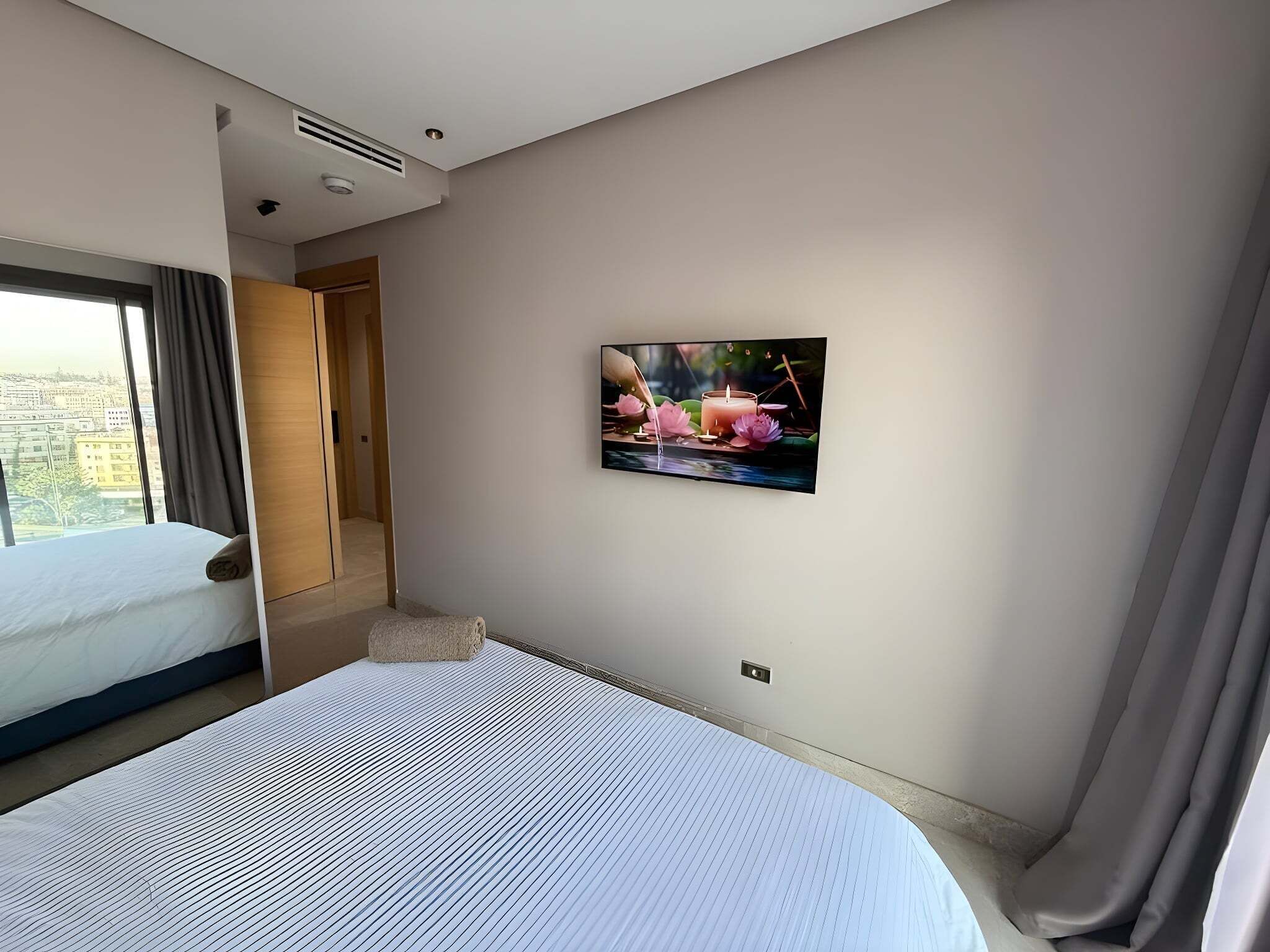 Ein Hotelzimmer mit Bett, einem Fenster mit Vorhängen, einem an der Wand befestigten Fernseher und einer Tür zu einem anderen Raum.