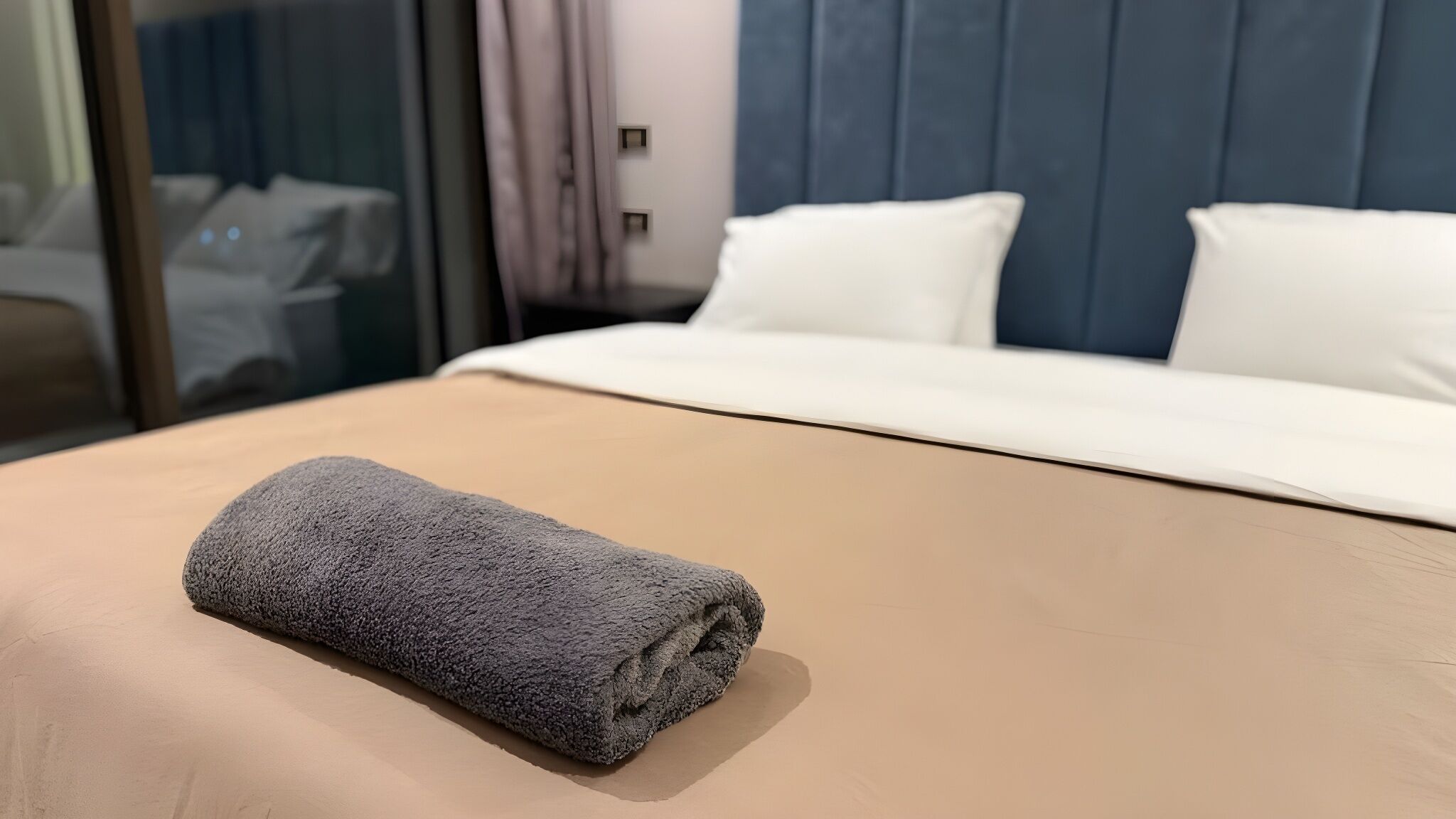 Ein ordentlich bezogenes Bett mit einem zusammengerollten Handtuch im Vordergrund, einem weißen Kopfkissen und einem blauen Kopfteil.
