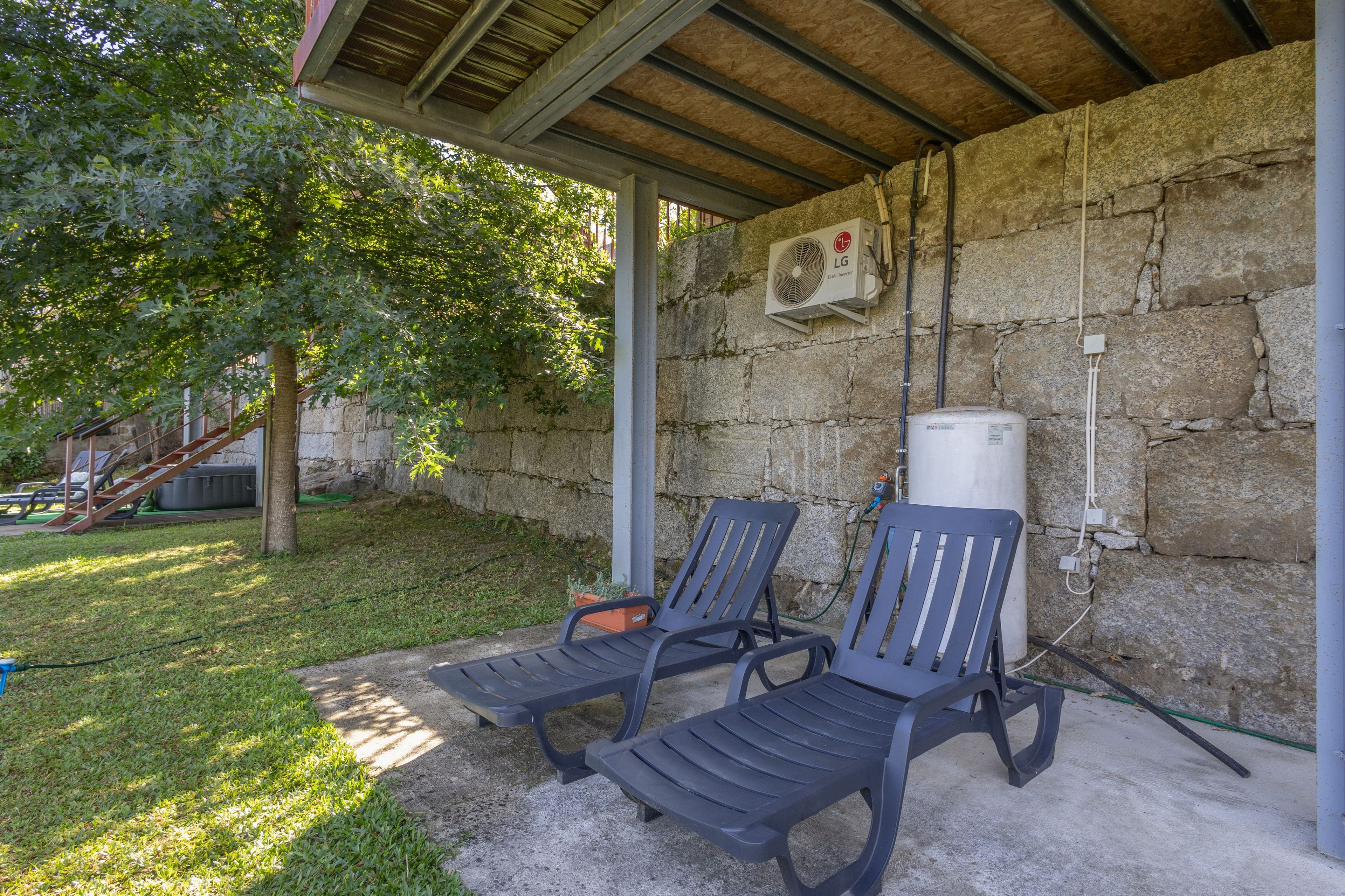 Un espace extérieur aménagé avec deux fauteuils confortables placés sous une pergola en bois, adossé à un mur en pierre, et équipé d’un climatiseur fixé au mur.