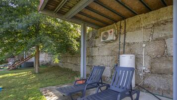 Un espace extérieur aménagé avec deux fauteuils confortables placés sous une pergola en bois, adossé à un mur en pierre, et équipé d’un climatiseur fixé au mur.