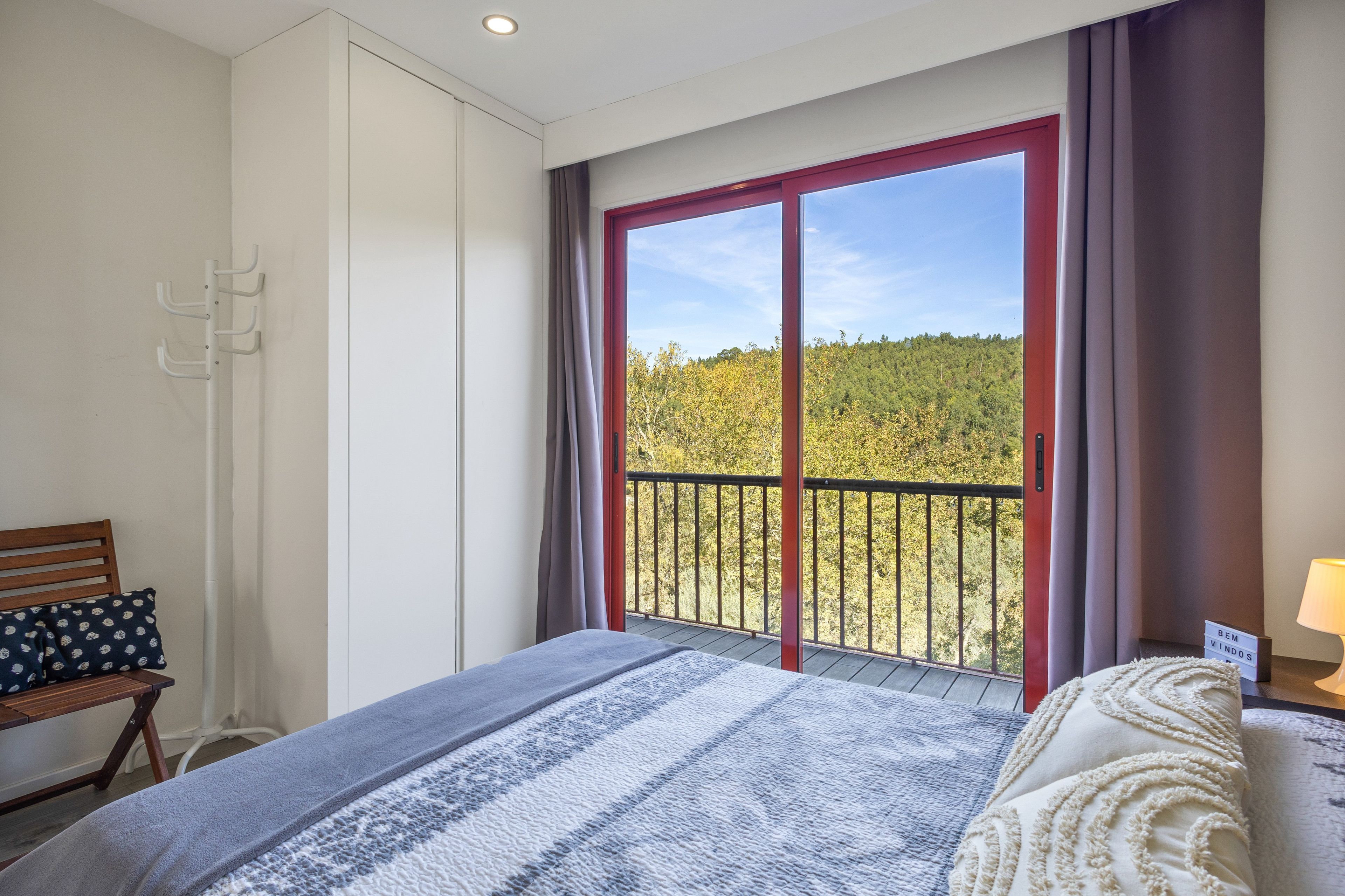Une chambre avec une grande fenêtre, un lit recouvert d’une couverture à rayures, un banc en bois et une petite table avec une lampe.