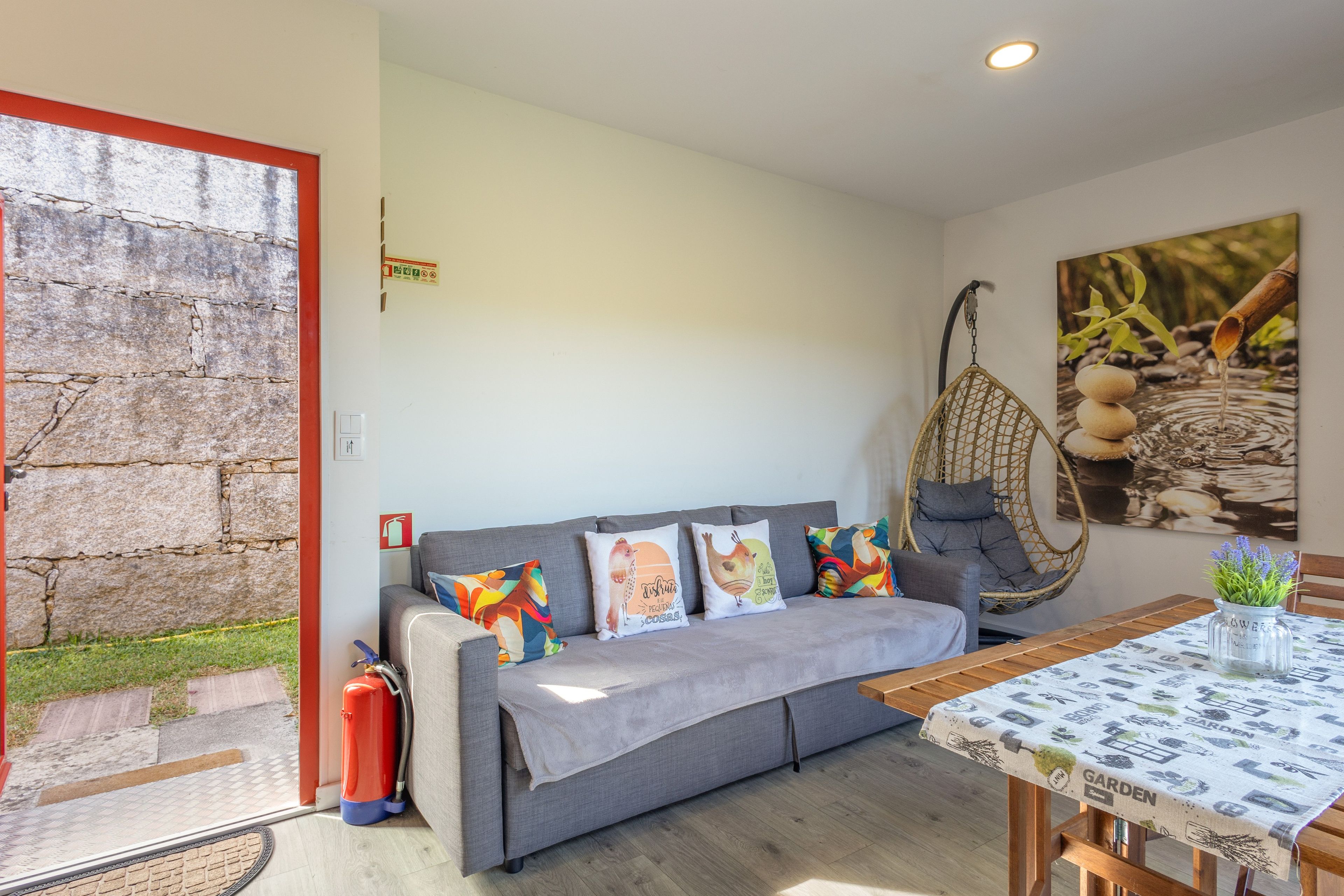 A cozy room with a grey sofa, colorful cushions, a hanging chair, a table with a garden-themed tablecloth, and a fire extinguisher near the door.