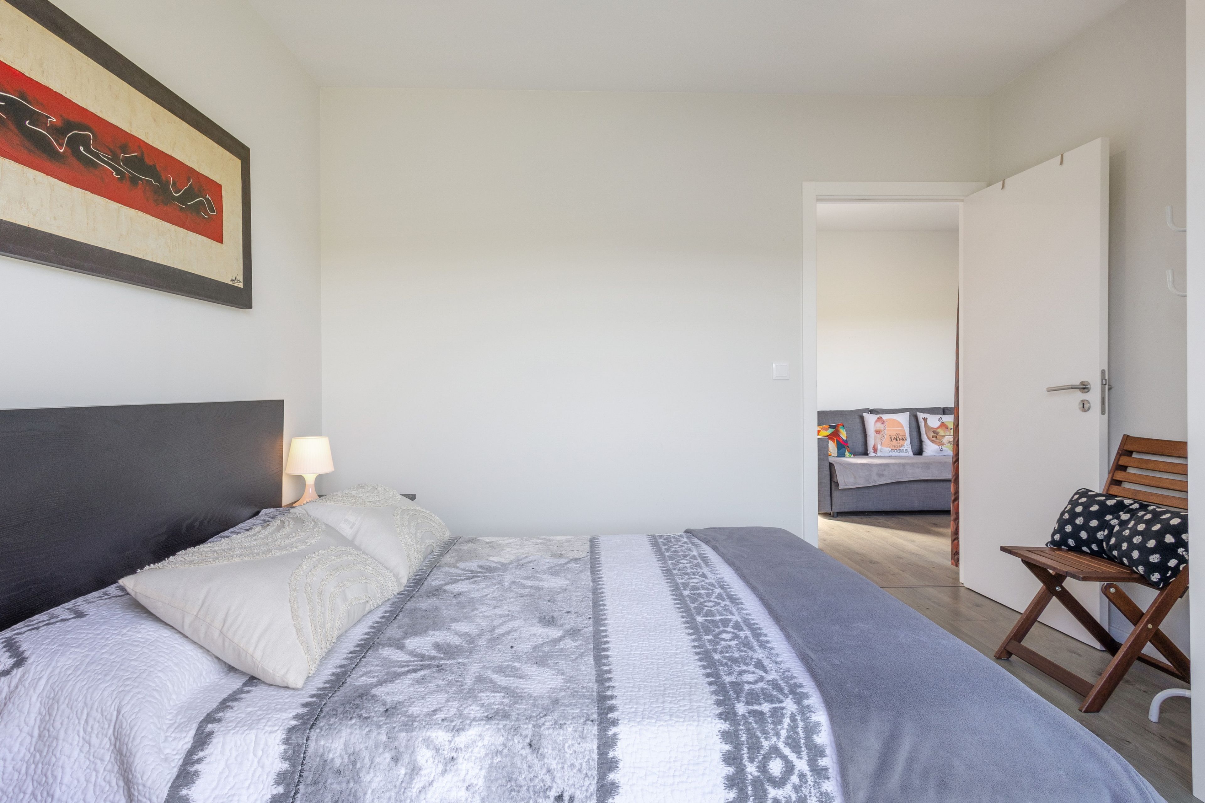A bedroom with a bed, a nightstand, a lamp, a chair, and a door leading to another room.