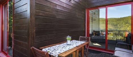 A wooden deck with a table and chairs, a glass door leading to a balcony, and a view of a forested hillside.