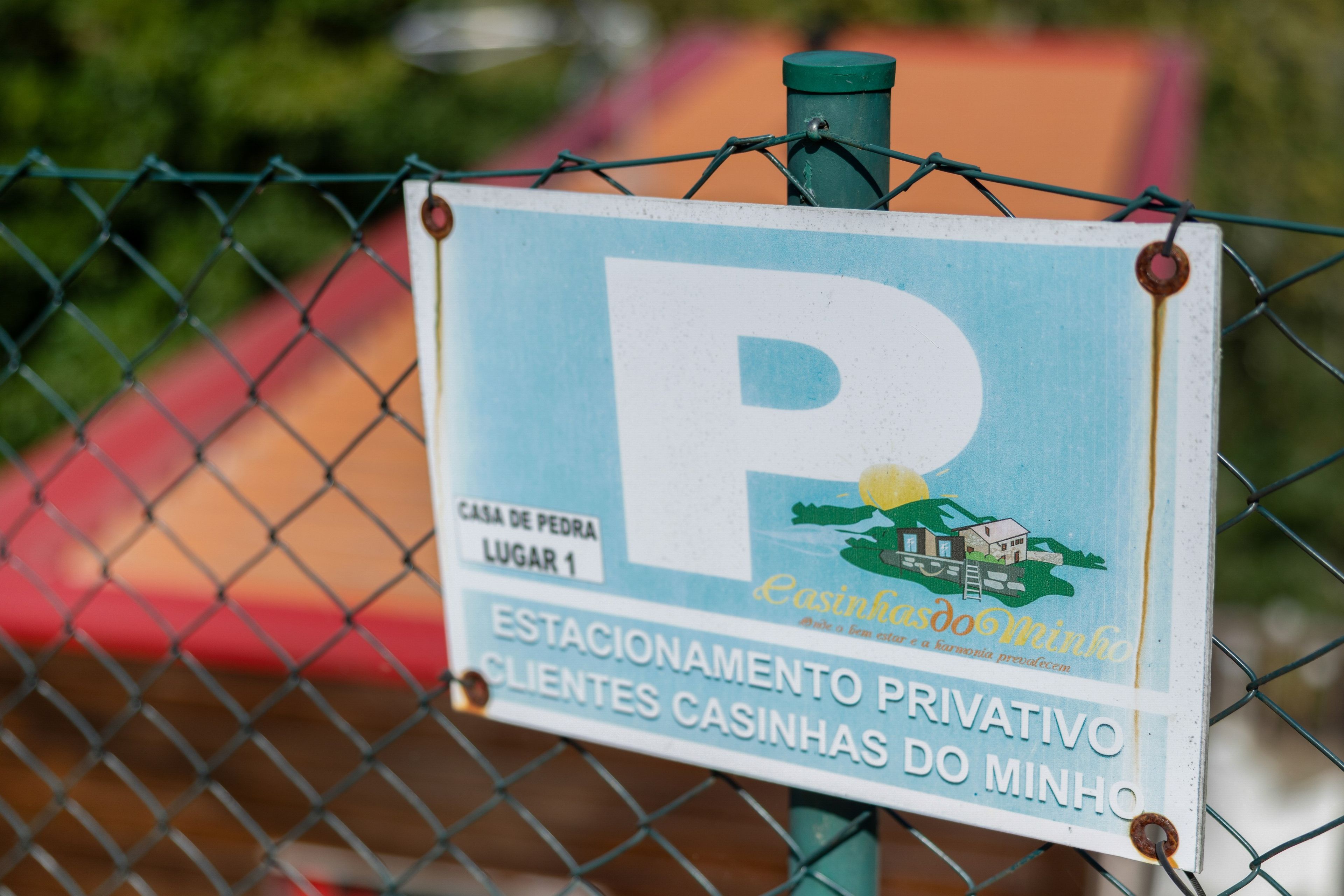Un panneau avec un grand « P » indiquant un espace de stationnement, un marqueur de localisation indiquant « CASA DE PEDRA LUGAR 1 » et une zone de stationnement privée intitulée « ESTACIONAMENTO PRIVATIVO  » réservée aux « CLIENTES CASINHAS DO MINHO ».