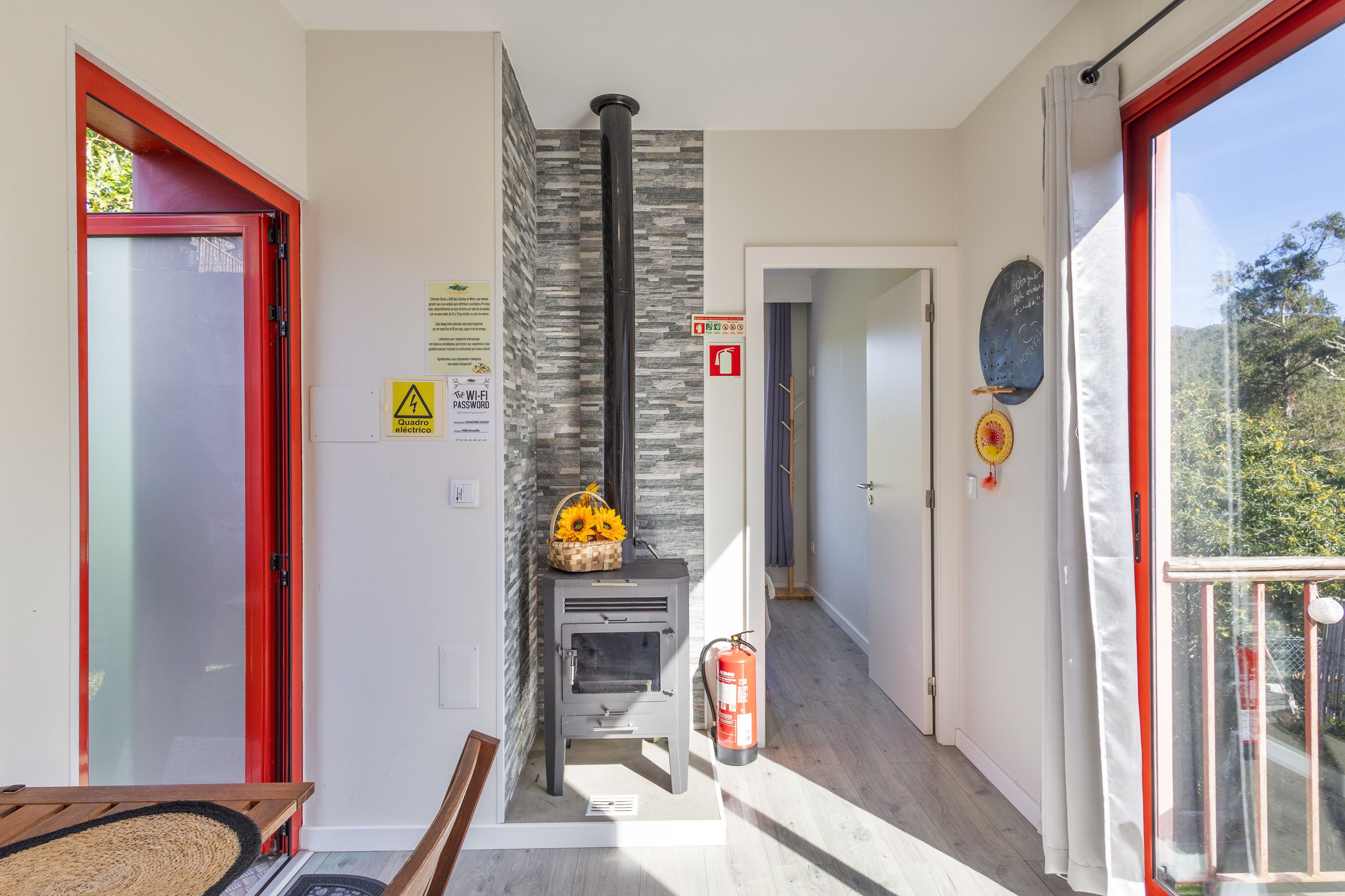 Una habitación con chimenea, una puerta con marco rojo, una pared con letrero de seguridad contra incendios y una ventana con vistas a árboles.