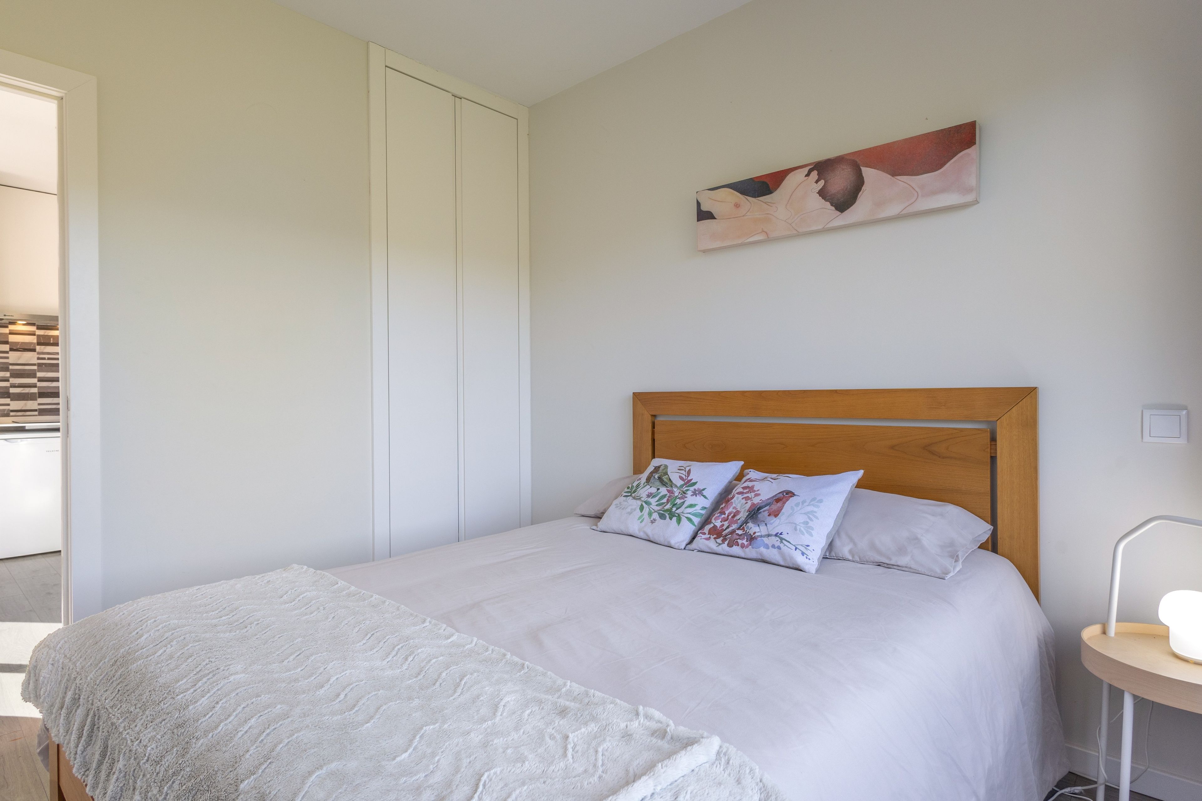 Un dormitorio con cama, almohadas, una colcha, cabecera de madera, un cuadro en la pared y una mesita de noche con lámpara.