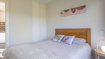 Un dormitorio con cama, almohadas, una colcha, cabecera de madera, un cuadro en la pared y una mesita de noche con lámpara.
