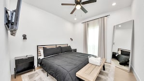 Ein modernes Schlafzimmer mit Bett, Nachttisch, an der Wand montiertem Fernseher, Deckenventilator und einem großen Spiegel.