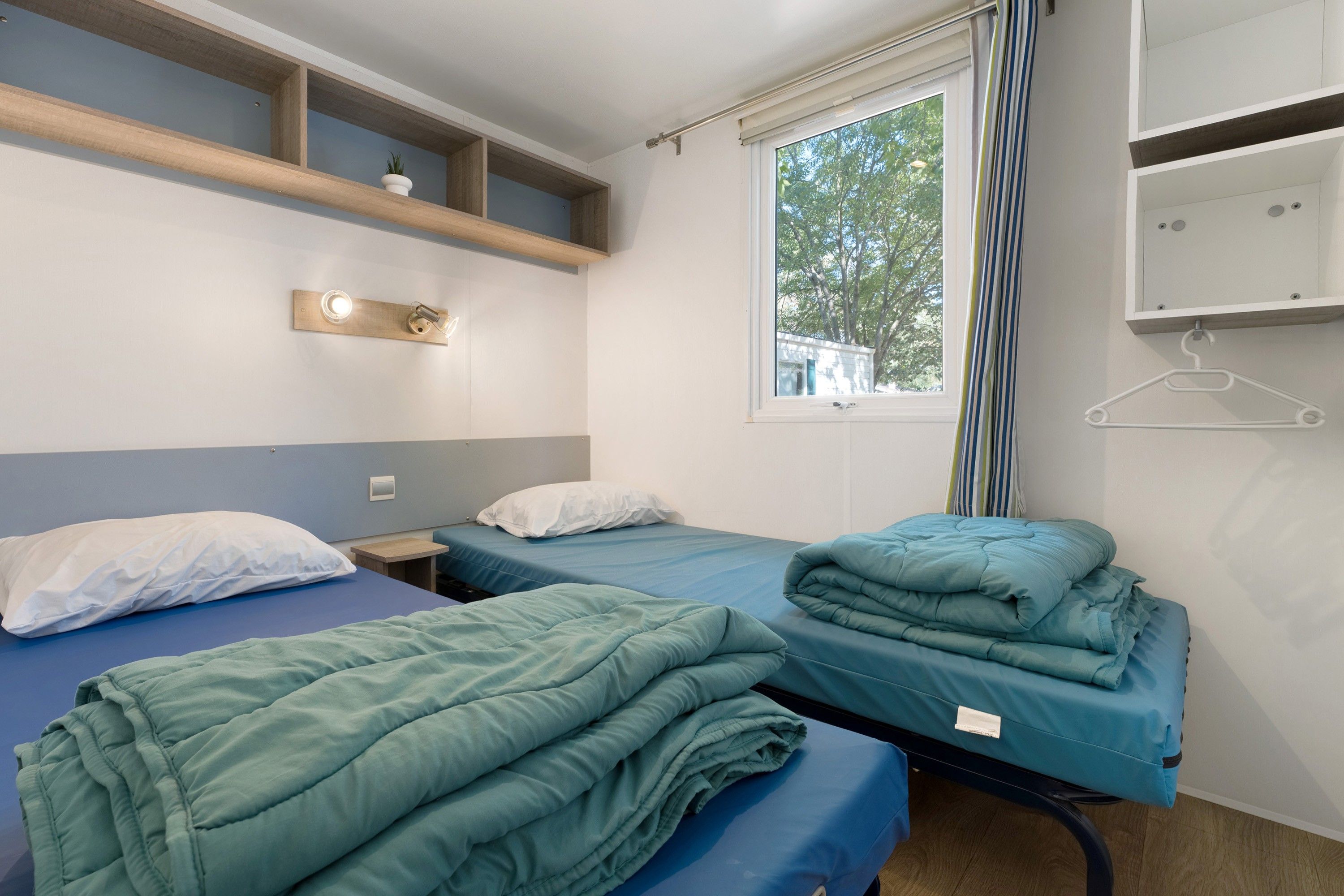 Ein Zimmer mit zwei Einzelbetten, einem kleinen Tisch, einem Regal mit einer Pflanze und einem Fenster mit Vorhängen.