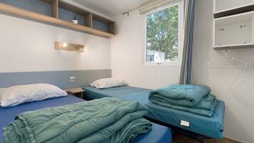Ein Zimmer mit zwei Einzelbetten, einem kleinen Tisch, einem Regal mit einer Pflanze und einem Fenster mit Vorhängen.