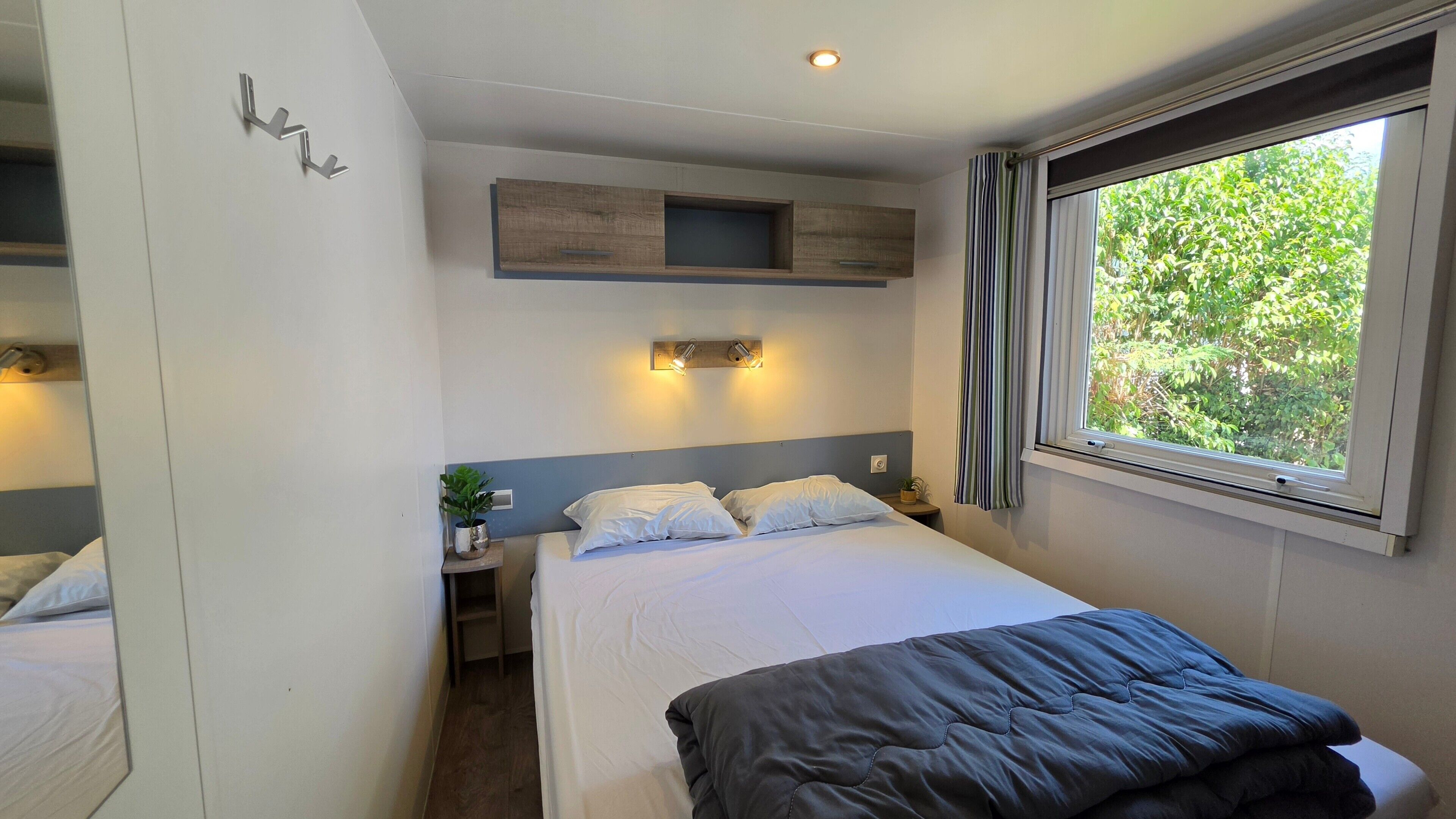 Ett hotellrum med en säng, sängbord, en liten hylla, ett fönster med gardiner och en väggmonterad lampa.