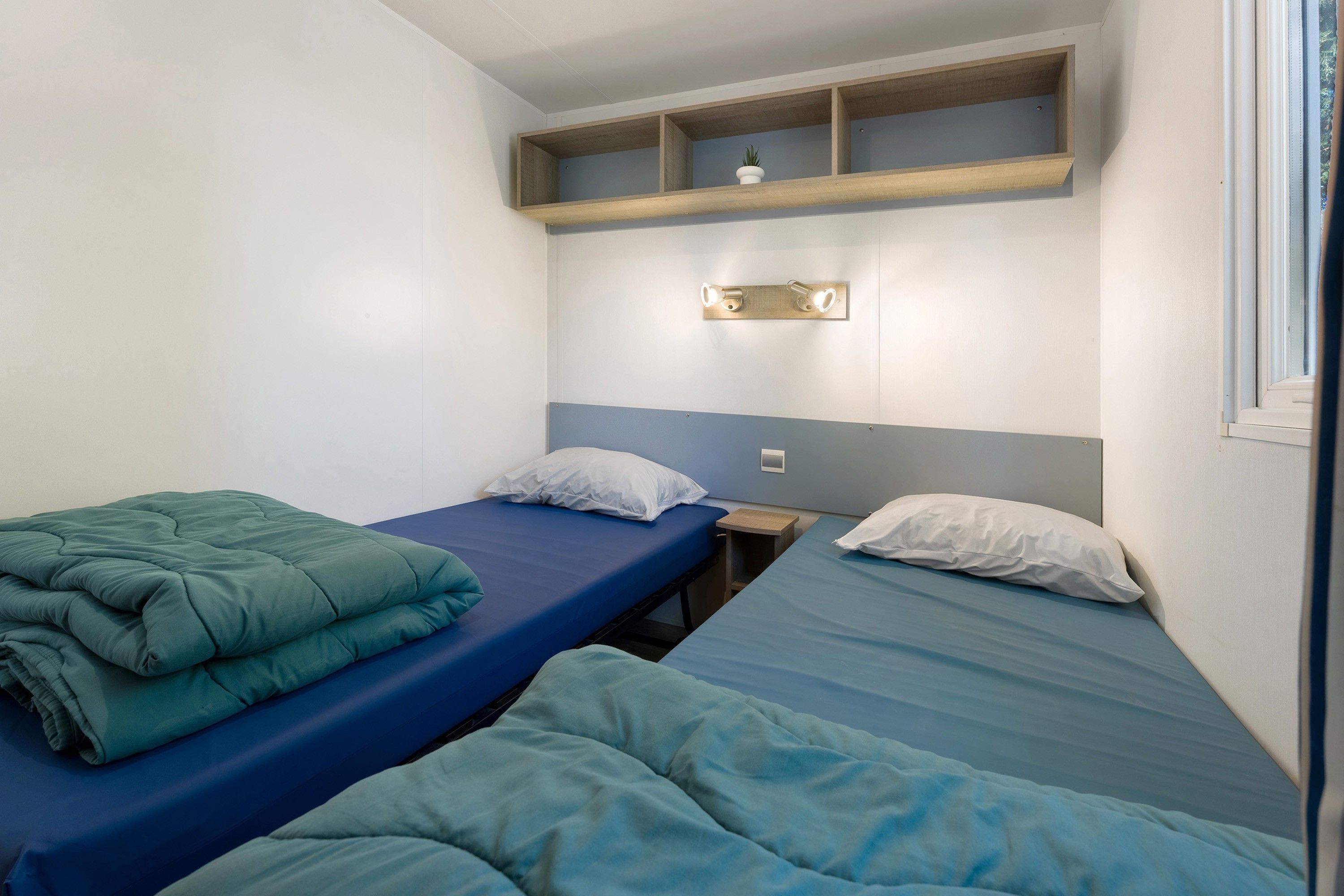A room with two beds, each with a blue mattress and white pillows, and a wooden shelf above the beds.