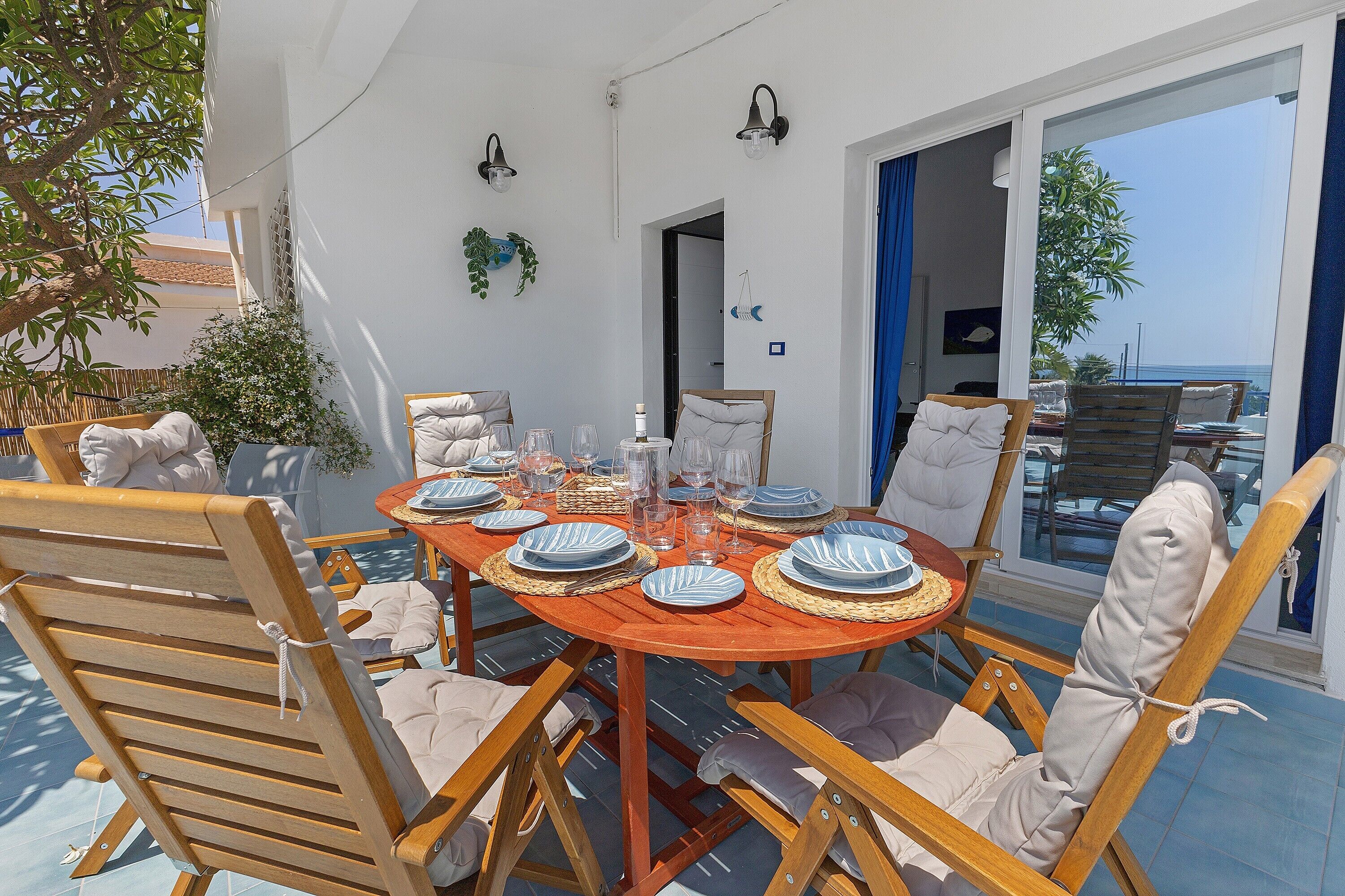 An outdoor dining area with a round table set for six, surrounded by wooden chairs with cushions.