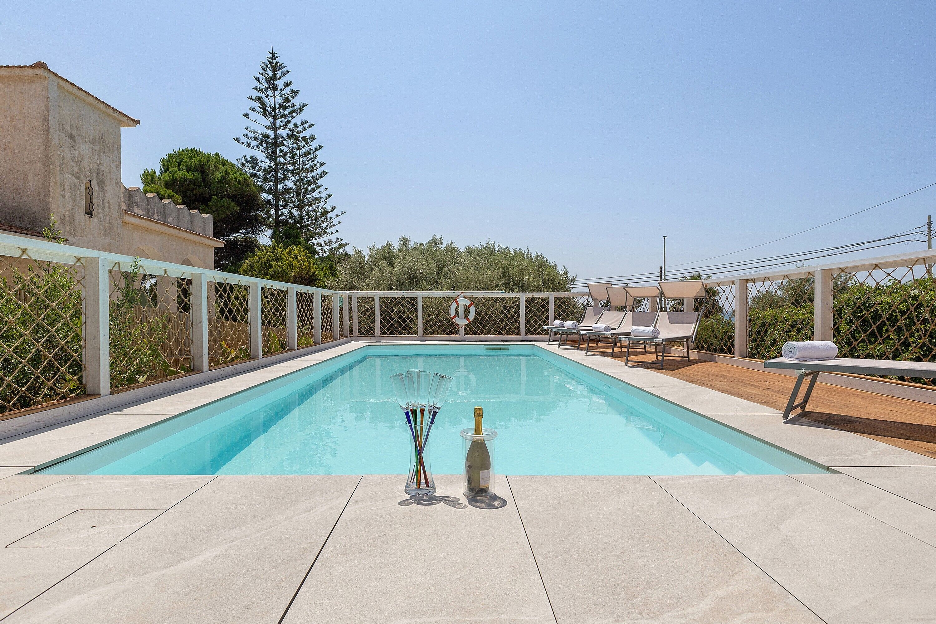 Una piscina rectangular al aire libre con superficie de agua azul cristalina, rodeada por una cerca blanca. Hay sillones de descanso y una pérgola con sombrillas.