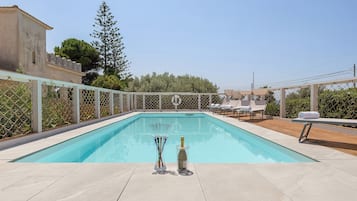Una piscina rectangular al aire libre con superficie de agua azul cristalina, rodeada por una cerca blanca. Hay sillones de descanso y una pérgola con sombrillas.