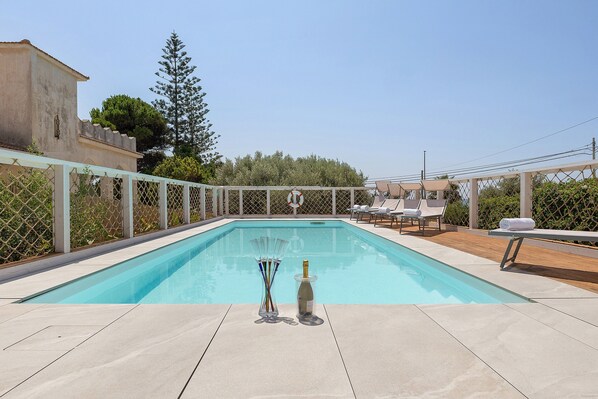 A rectangular outdoor swimming pool with a clear blue water surface, surrounded by a white fence. There are lounge chairs and a pergola structure with umbrellas.