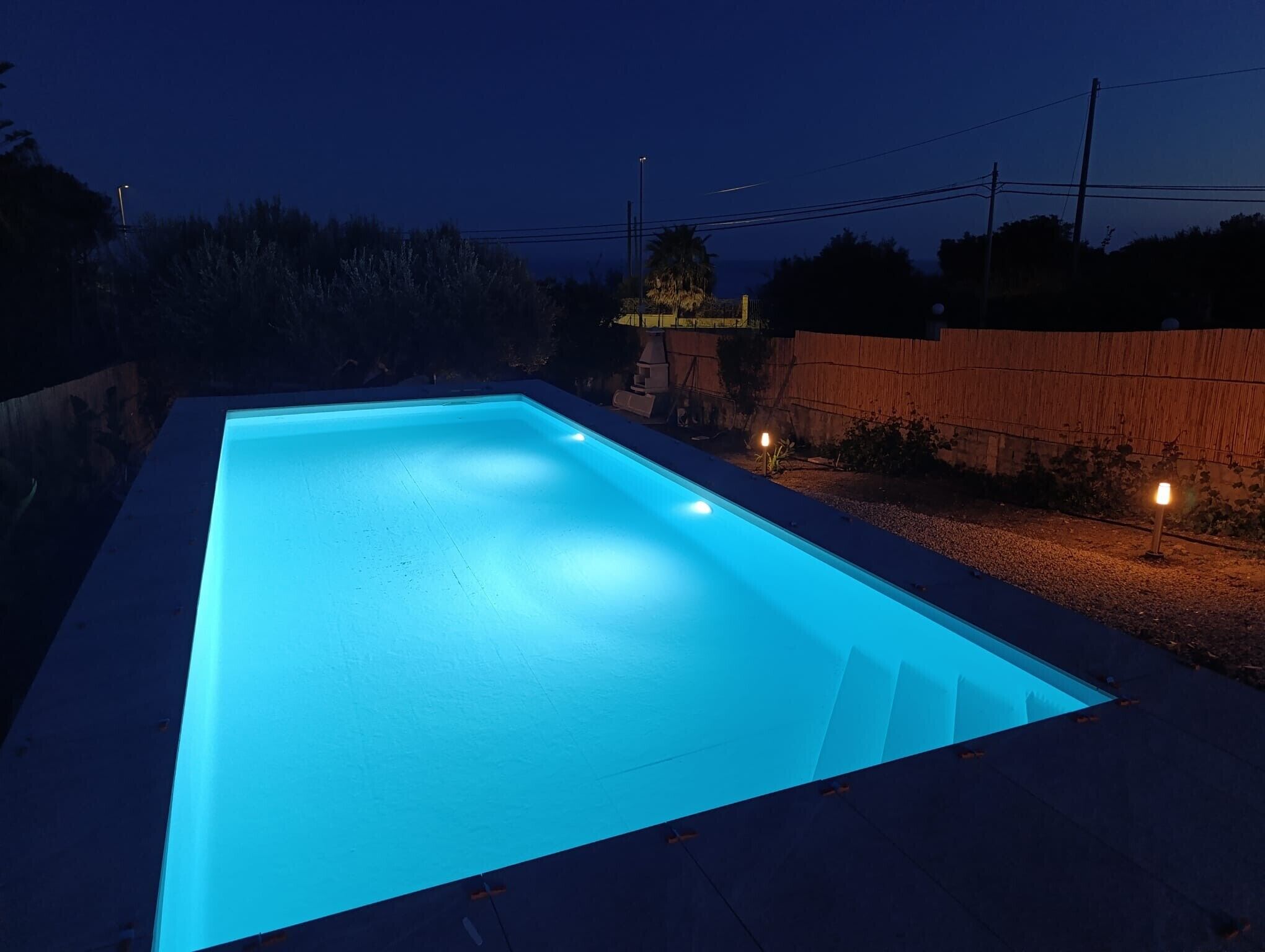Una piscina rettangolare con interni illuminati di blu, circondata da una recinzione in legno e un'area ghiaiosa.