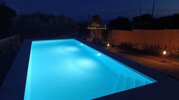 Una piscina rettangolare con interni illuminati di blu, circondata da una recinzione in legno e un'area ghiaiosa.