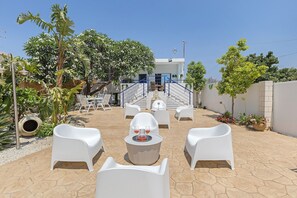 An outdoor seating area with white chairs and a table, surrounded by greenery and a building with a staircase.