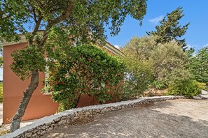 A paved courtyard with a stone wall, a tree with green foliage, and a building with a reddish-brown facade. - Holiday Home 'Villa Meltemi' with Sea View, Wi-Fi, and Air Conditioning (Tsoukalades)