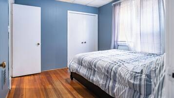 Una camera da letto con pavimento in legno, un letto con lenzuola a righe, due porte bianche e una finestra con tende leggere.