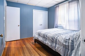 Een slaapkamer met een houten vloer, een bed met gestreepte lakens, twee witte deuren en een raam met dunne gordijnen.